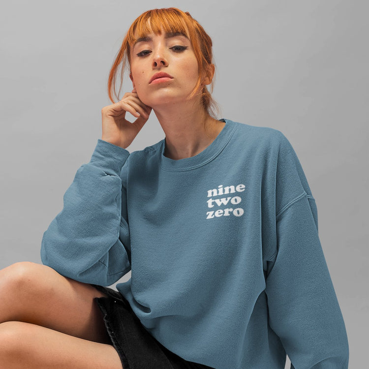 red haired woman with a blue baggy crewneck sweatshirt with the text nine two zero for the area code