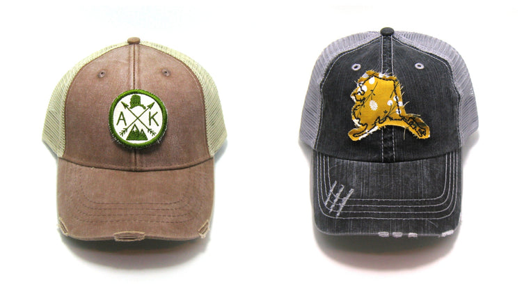 Alaska Trucker hat collage one brown hat with olive green and cream arrow patch and one gray distressed trucker hat with a mustard yellow fabric patch in the shape of alaska