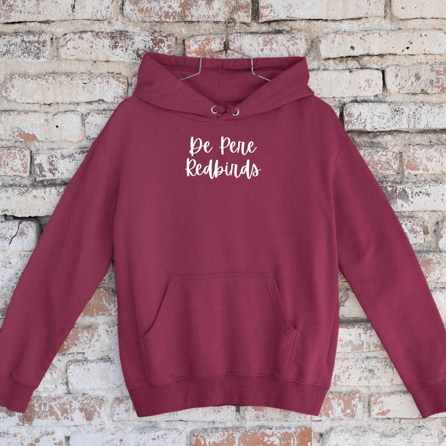 cardinal red hoodie sweatshirt with script "de pere redbirds" on center chest, hanging on hanger in front of brick wall